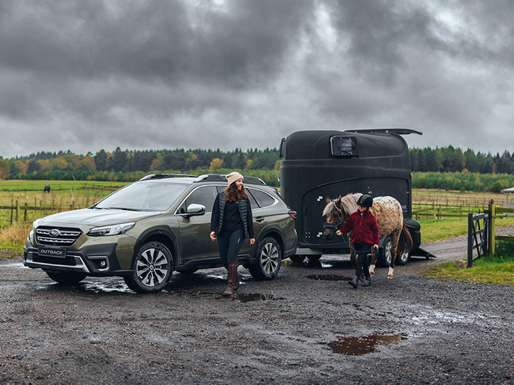 Subaru Outback ar zirgu piekabi dubļos, kamēr sieviete un meitene iet blakus – meitene ved zirgu.